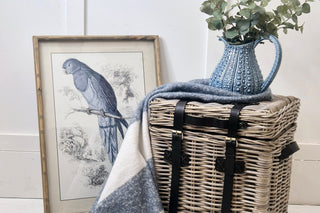 Wicker picnic basket with black straps, blue and white plaid blanket, and blue pitcher with eucalyptus on a white surface.