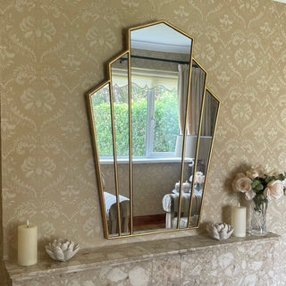 Gresham Mirror, arched geometric wall mirror with a gold frame hangs above a marble fireplace mantel decorated with candles and flowers, set against patterned beige wallpaper. Mirror by www.lilybloom.ie