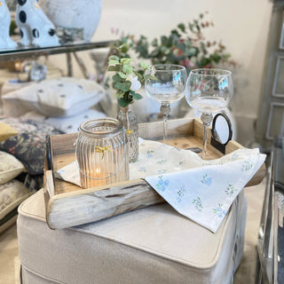 Decorative tray with glasses, candle, and napkins on a gray ottoman in a living room.
