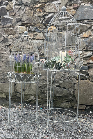 Two decorative metal birdcage plant stands against a stone wall.