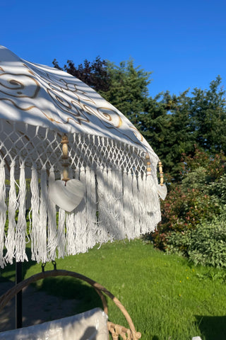 A white bohemian-style outdoor umbrella with intricate gold embroidery and long fringe tassels, set in a grassy garden under a clear blue sky.