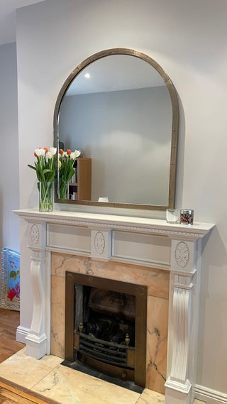 An arched wall mirror with a distressed bronze frame hanging above a white fireplace mantel