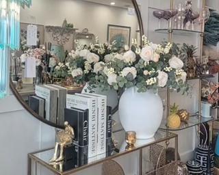 Decorative interior setting with a round mirror, books, and floral arrangements.