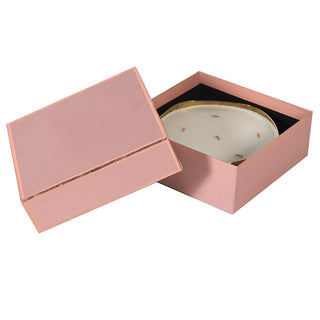 A square pink gift box with its lid off, displaying a white oval dish with a gold rim and small brown botanical inclusions on a black insert, set against a plain white background.